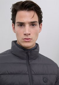 Szara ocieplana kurtka z wysokim kołnierzem, zamkiem błyskawicznym z przodu oraz okrągłym logo na lewym obszarze klatki piersiowej. Gładka tekstura i pikowany wzór.