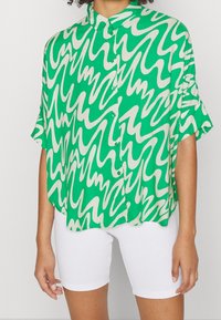 Grön kortärmad skjorta med ett vågigt vitt mönster, knäppning med knappar och krage, tillsammans med vita shorts. Slät tygtextur.