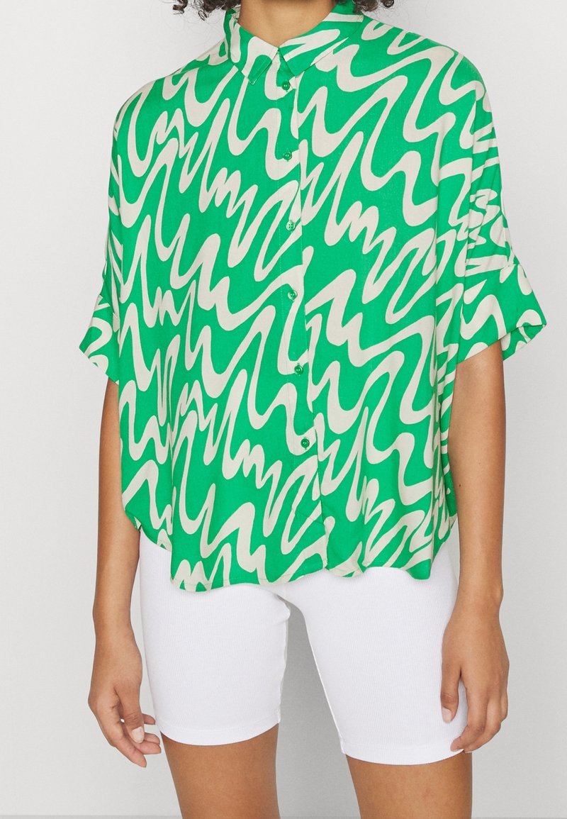 Grön kortärmad skjorta med ett vågigt vitt mönster, knäppning med knappar och krage, tillsammans med vita shorts. Slät tygtextur.