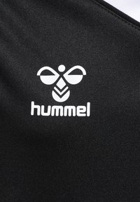 Czarna koszulka z białym logo o wzorze inspirowanym pszczołami oraz słowem "hummel" napisanym pogrubioną czcionką poniżej. Gładki, teksturowany materiał.