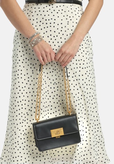 Melna ādas rokas soma ar zelta metāla aizdari un ķēdes siksnu, ko tur persona polka punktu kleitā un ar pērlītēm rotātām aprocēm.