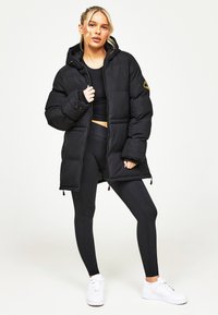 Melns polsterēts mētelis ar kapuci, ar rāvējslēdzēju priekšā, elastīgām aprocēm un sānu kabatām ar rāvējslēdzēju. Nēsāts virs melnām legingām un baltajiem sporta apaviem.