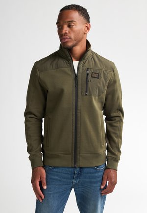 Olijfgroene zip-up jack in een gestructureerde stof met een hoge kraag, voorzien van een borstzak en zijzakken, gedragen over een blauwe spijkerbroek.