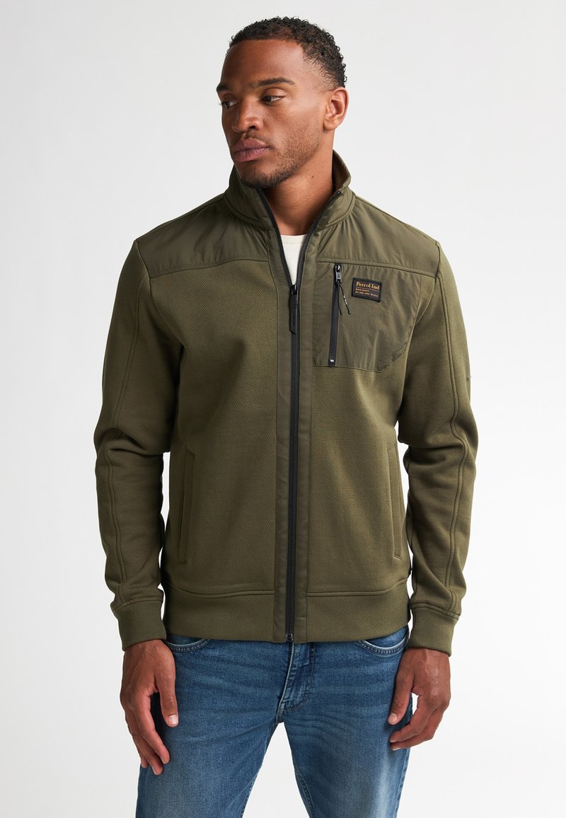 Olijfgroene zip-up jack in een gestructureerde stof met een hoge kraag, voorzien van een borstzak en zijzakken, gedragen over een blauwe spijkerbroek.