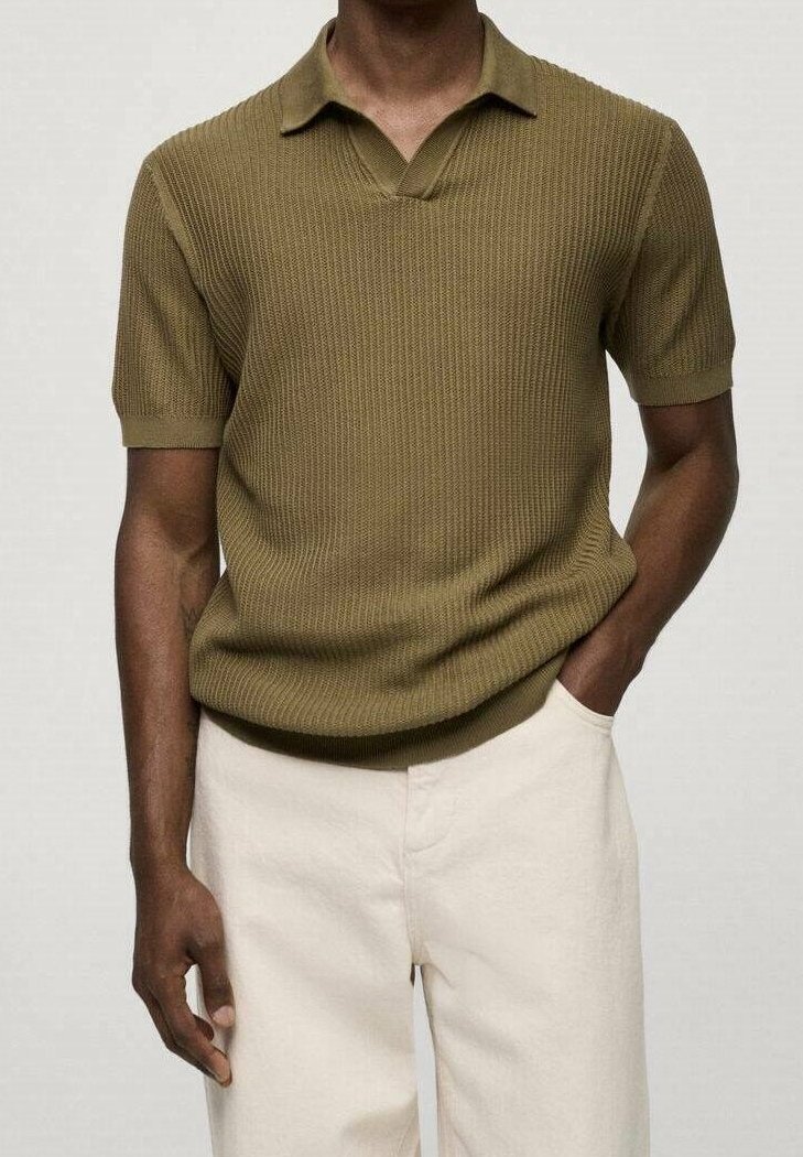 Homme portant un polo en maille vert olive à manches courtes, légèrement rentré dans un pantalon blanc cassé, avec une main dans la poche.