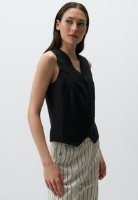 Gilet en lin noir avec un décolleté en V et une fermeture à boutons, doté d'une silhouette ajustée et d'un ourlet court. Associé à un pantalon léger à rayures.