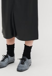 Saia de couro preta com bainha recortada, combinada com sapatilhas desportivas cinzentas e meias pretas com efeito canelado, apresentando uma borda superior ondulada.