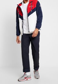 Windbreaker jacket featuring red, navy, and white panels, zippered front, elastic cuffs, worn over a white t-shirt and navy pants with rolled cuffs.