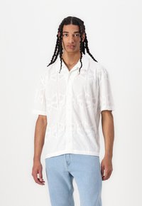 Une chemise à manches courtes blanche avec des boutons qui présente un motif géométrique subtil. La chemise a un col et est fabriquée dans un tissu léger et respirant.
