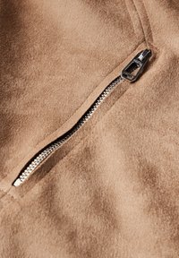 Acercamiento a un zipper plateado sobre una tela de gamuza en color beige, mostrando la textura y los detalles de costura alrededor del área del zipper.