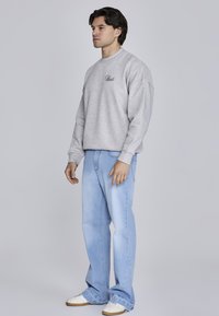 Pelēks sporta krekls ar noapaļotu kakla izgriezumu un melnu izsistu logotipu, apvienots ar gaiši ziliem taisnajiem džinsiem un baltiem sporta apaviem.