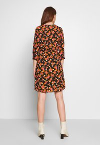 Kvinna klädd i en knälång svart klänning med orange och rosa blommönster, stående med ryggen mot oss, kombinerat med vita ankelstövlar.