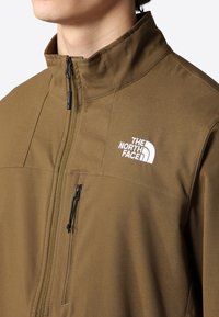 Brun lynlåstrøje med høj krave, med en hvid logo på brystet og en sidelomme med lynlås. Glat, tekstureret stof.