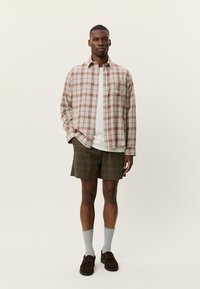 Καρό πουκάμισο με κουμπιά σε μπεζ και μπορντό, φορεμένο πάνω από λευκό T-shirt· συνδυασμένο με χακί σορτς με σχέδια και καφέ μοκασίνια.