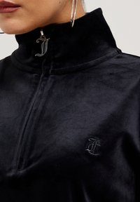 Close-up van een persoon die een zwarte fluwelen rits-top met hoge kraag en zilveren ringoorbellen draagt, waarbij een ritssluiting en een geborduurd logo zichtbaar zijn.