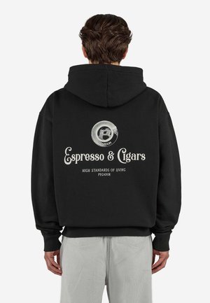 Person iført en sort hættetrøje med en kaffekop-logo og teksten "Espresso & Cigars, High Standards of Living, Pegador" på ryggen.