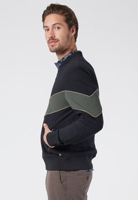 Felpa blu navy con pannello diagonale verde, piping crema, tasche con zip, orlo e polsini a costine. Indossata sopra una camicia a fiori con colletto.