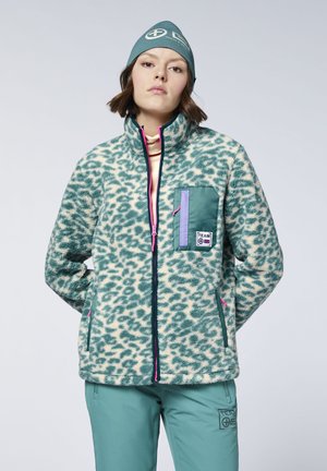 Young person wearing teal patterned fleece jacket with pink zipper, matching teal pants and beanie, standing with hands behind back.