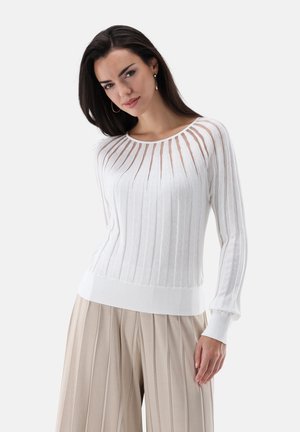 Frau mit dunklen Haaren, die einen weißen, gerippten Pullover mit langen Ärmeln und beige plissierte, weit geschnittene Hosen trägt, stehend vor einem einfarbigen Hintergrund.