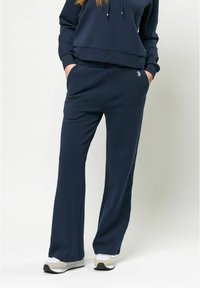 Navy krāsas sporta bikses atvieglotā piegriezumā, izgatavotas no mīksta auduma, ar sānu kabatām un izšūtu logotipa detaļu. Apvienotas ar atbilstošu jaku.