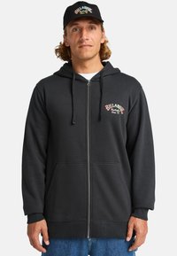 Zwarte zip-up hoodie met voorkleppen en geribbelde manchetten. Beschikt over geborduurde tekst "Billabong Australia Since '73" in kleurrijke letters.