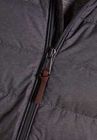 Grå puffjacka med slät textur, med en sidodragkedja och en dragflik i brunt läder. Sömmarna synliga på tyget.