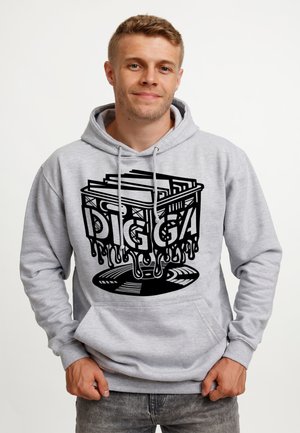 Młody mężczyzna w jasnoszarej bluzie z kapturem z czarną grafiką ściekającej skrzyni wypełnionej płytami i napisem "DIGGA" z przodu.