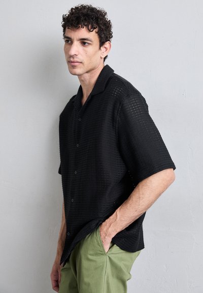 Chemise noire à manches courtes avec un design en tricot texturé et ajouré. Portée sur un pantalon vert, avec une coupe décontractée et un col.