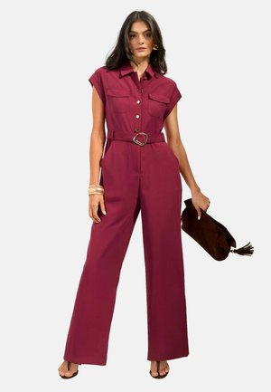 Vrouw gekleed in een mouwloze bordeauxrode jumpsuit met wijde pijpen en een ceintuur, houdt een bruine clutch vast en draagt armbanden en sandalen.