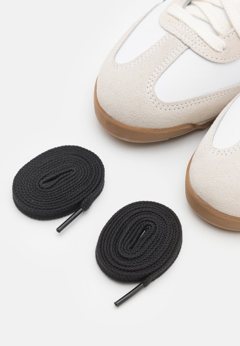 Baskets en daim et cuir de couleur beige clair et blanche, avec une semelle en caoutchouc naturel. Deux lacets noirs enroulés à côté des chaussures.