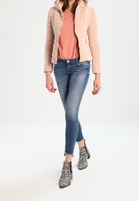 Chaqueta acolchada de color rosa claro con capucha, combinada con una camiseta rosa y unos pantalones ajustados de mezclilla azul deslavado. Botas de tobillo de estampado animal en blanco y negro.