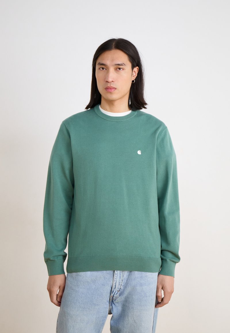Green crewneck sweater in soft fabric, featuring long sleeves and a small white logo on the chest. Paired with light blue jeans.