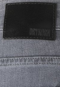 Harmaa kangas hienovaraisella tekstuurilla, jossa on mustasta nahasta valmistettu etiketti, jossa on kohokuvioituna "DRYKORN." Näkyvät ompeleet reunoilla.