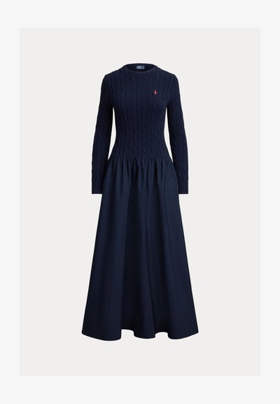 Σκουρό μπλε φόρεμα με πλεκτό πάνω μέρος σε σχέδιο καλωδίου και μακριά μανίκια, με φούστα σε σχήμα φλογέρας. Κόκκινη λεπτομέρεια λογότυπου στο στήθος. Ομαλή υφή σε όλο το φόρεμα.