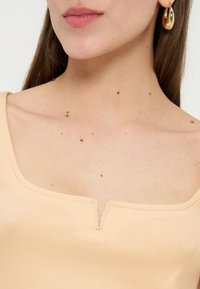 Το κάτω μέρος του γυναικείου προσώπου και ο λαιμός με πολλαπλές καστανές ελιές, φοράει μπεζ τοπ με τετράγωνο ντεκολτέ και χρυσούς κρίκους στα αυτιά.