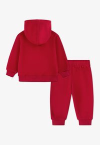 Felpa rossa con cappuccio e polsini a coste, abbinata a pantaloni della tuta rossi con fascia elastica in vita e polsini affusolati.