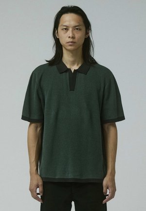 Giovane uomo con capelli lunghi e neri che indossa una polo a maniche corte di maglia verde scuro con collo e rifiniture nere, in piedi davanti a uno sfondo uniforme.