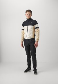 Giacca da track nera, crema e beige con strisce bianche; polsini elastici. Pantaloni neri abbinati con orlo elastico, indossati con sneakers nere.