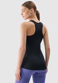 Czarny sportowy top na ramiączkach z wycięciem na plecach, o dopasowanej sylwetce i gładkiej fakturze. W zestawie z fioletowymi spodniami do jogi.