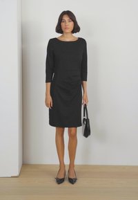 Abito nero fino al ginocchio con maniche a tre quarti e una silhouette aderente. Abbinato a tacchi alti neri e una piccola borsa. Tessuto liscio.