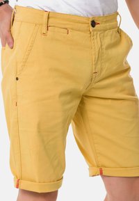 Gelbe knielange Shorts mit umgeschlagenen Säumen, ausgestattet mit zwei Vordertaschen, einem Knopfverschluss und roten Nähten. Hergestellt aus einem glatten Stoff.