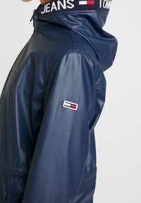 Navy blue vesimproof takki, jossa on vetoketju edessä ja huppu. Ominaisuuksiin kuuluu kirjailtu logo-ompelu hihassa ja joustava vyötärö.