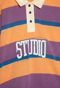 Lilla og oransje stripete polo-skjorte med hvit krage, med et frontlogo som leser "STUDIO" i fete, blå bokstaver.