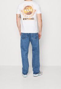 Camiseta blanca con un gráfico colorido de NASA y Da Vinci impreso en la parte posterior, combinada con jeans de corte holgado y zapatillas multicolores.