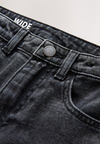 Primo piano della vita di jeans in denim nero con passanti per cintura, bottone di metallo inciso "Next Authentic Since 1992" e etichetta bianca "WIDE" all'interno.