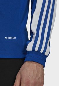 Maillot de sport bleu à manches longues avec des rayures blanches sur les manches, un tissu texturé, et un logo "AEROREADY" sur l'ourlet.