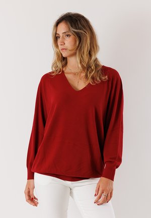 Maglione rosso a V con maniche lunghe e ampie, polsini a coste e una vestibilità rilassata. Realizzato in un tessuto liscio e morbido. Indossato con pantaloni bianchi.
