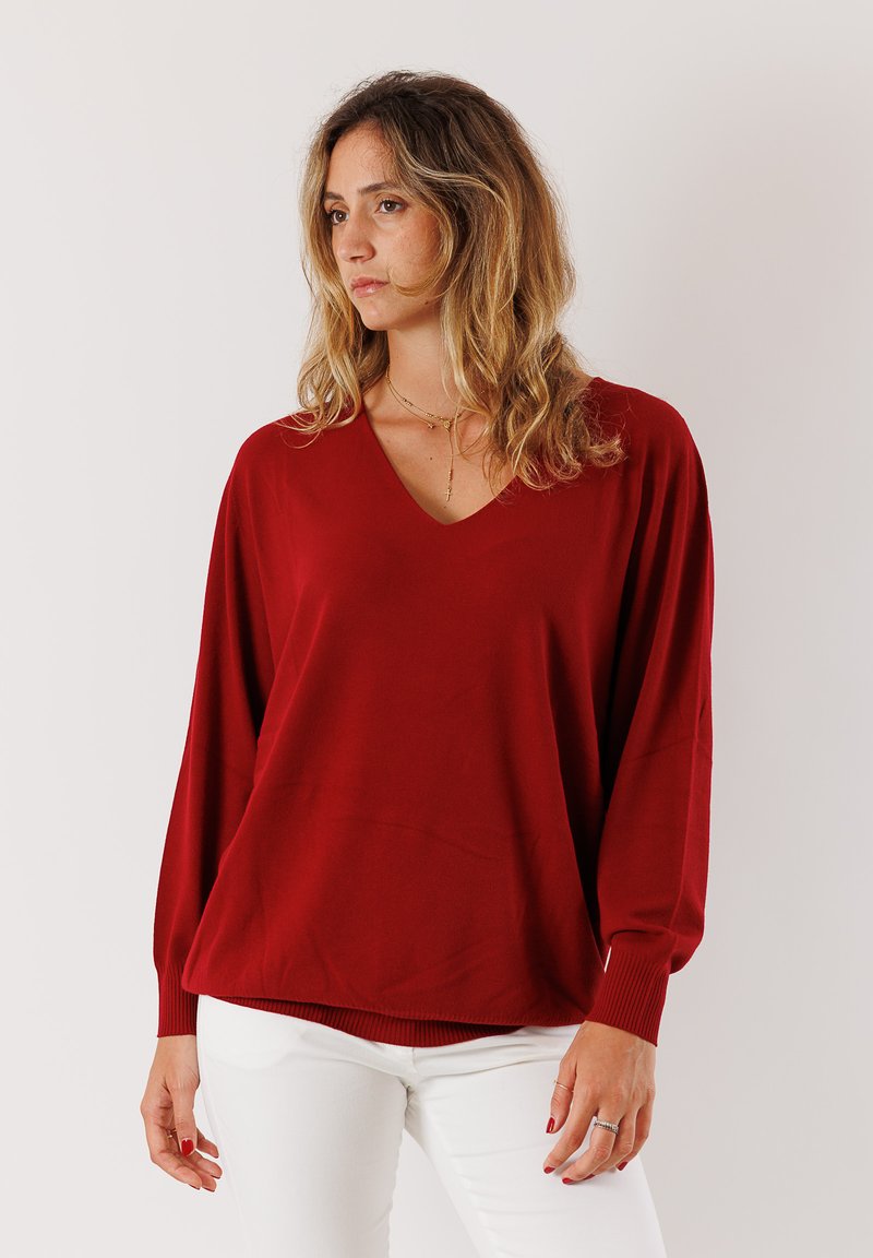 Maglione rosso a V con maniche lunghe e ampie, polsini a coste e una vestibilità rilassata. Realizzato in un tessuto liscio e morbido. Indossato con pantaloni bianchi.