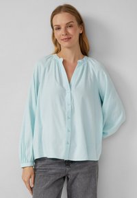 Donna che indossa una camicia con colletto gather azzurro chiaro, con maniche lunghe e jeans grigi scuri, in piedi contro un muro neutro.