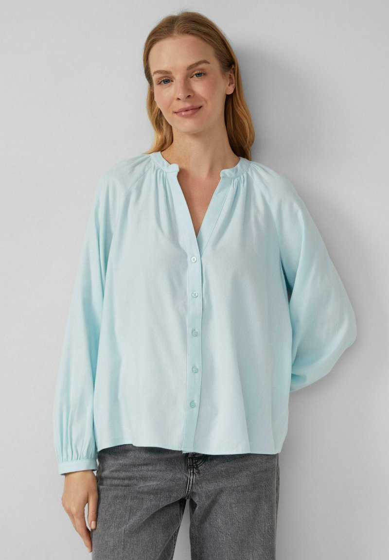 Donna che indossa una camicia con colletto gather azzurro chiaro, con maniche lunghe e jeans grigi scuri, in piedi contro un muro neutro.
